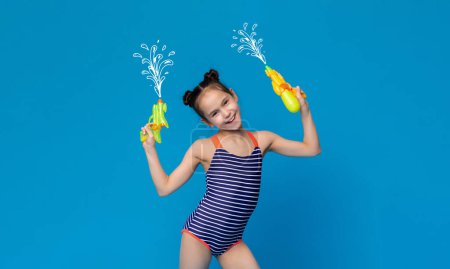 Summer fun toy. Little girl in swimsuit holding water guns, smiling on blue studio background
