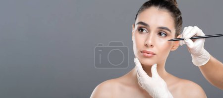 A young woman is receiving a cosmetic treatment in a clinic, where a professional is marking her face with a pencil while another hand holds her chin.