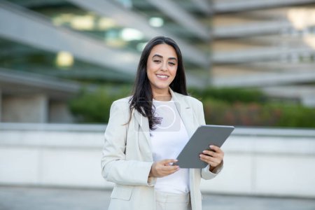 Une jeune femme d'affaires, vêtue d'un blazer blanc et d'un haut blanc, sourit en toute confiance alors qu'elle tient une tablette devant un bâtiment moderne. La scène dépeint un cadre professionnel