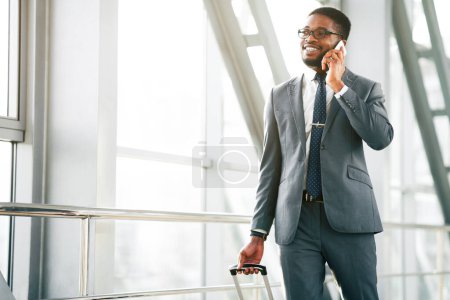 African American professional traveler talking on his phone outside a sleek airport, holding his luggage. The mood is focused and efficient, capturing the essence of a business trip, copy space