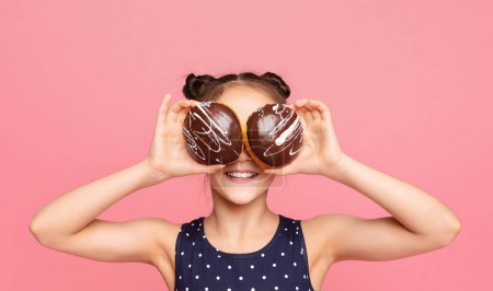 A cheerful young girl with a playful expression holds two chocolate donuts over her eyes against a vibrant pink background. She is wearing a polka dot dress.
