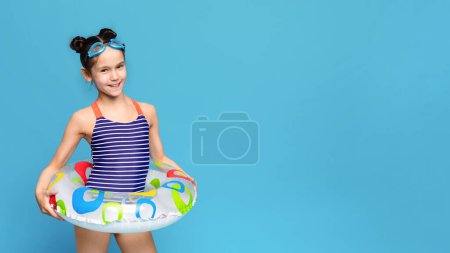 A young girl wearing a colorful bathing suit stands holding an inflatable pool float. The girls hair is tied up in a ponytail as she grips the float with a smile on her face, copy space