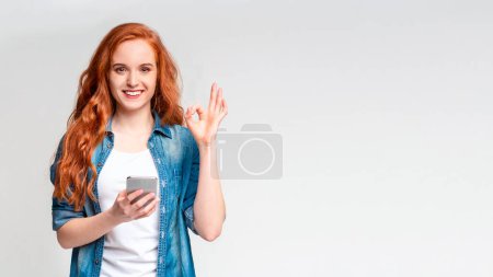 A cheerful young woman with long red hair is standing against a neutral grey backdrop, appears to be interacting with someone, making an OK sign with her hand while holding a smartphone in the other.