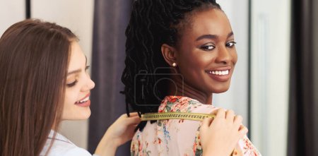 A professional tailor is capturing the measurements of a delighted young black woman for a custom dress tailoring session inside a well-lit modern studio