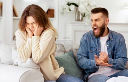 Couple Arguing. Husband Shouting To Desperate Wife, Sitting On Couch At Home