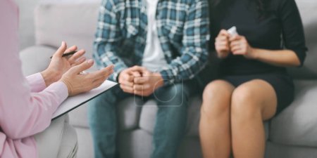 A therapist sits on a grey couch with a couple in front of them. The therapist is taking notes and listening to the couple. Therapist taking notes, possibly about the couples experiences or concerns.