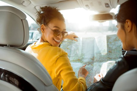 Eine junge schwarze Frau mit Sonnenbrille lächelt in die Kamera, während sie mit einer anderen Person auf dem Beifahrersitz über eine Landkarte blickt. Shes trägt einen gelben Pullover