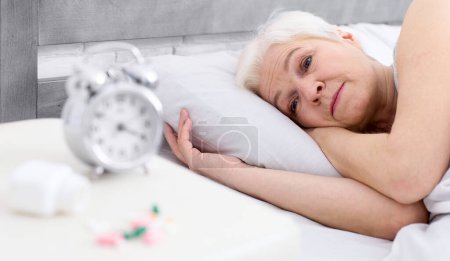 Photo for A woman is laying in bed, with her eyes open, next to an alarm clock set on the bedside table. The clock displays a time indicating the morning hour. - Royalty Free Image