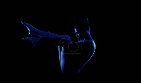 Woman posing in a dark room. Her legs are outstretched in a position that suggests she is either floating or leaping. The only source of light is a bright blue glow emanating from her body.