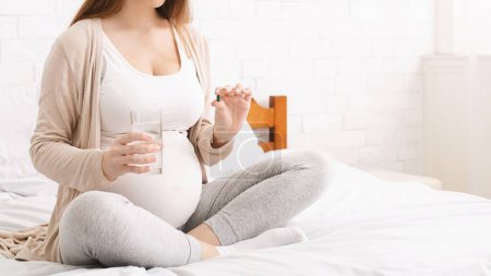 Eine schwangere Frau sitzt im Schneidersitz auf ihrem Bett und hält ein Glas Wasser und eine Tablette in einem hellen, sonnendurchfluteten Raum. Sie wirkt ruhig und entspannt in einer gemütlichen Umgebung, beschnitten, Kopierraum