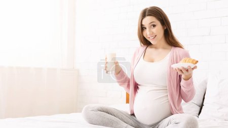 A pregnant woman is sitting comfortably in bed, smiling while holding a glass of milk and a pastry. She appears content and relaxed, showcasing a moment of calm and nourishment during her pregnancy.