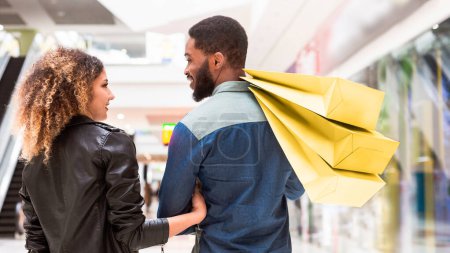 Afroamerikaner und Afroamerikanerinnen stehen in einem belebten Einkaufszentrum, halten mehrere Einkaufstüten in der Hand und genießen ihren gemeinsamen Einkaufsbummel durch verschiedene Geschäfte, Rückansicht