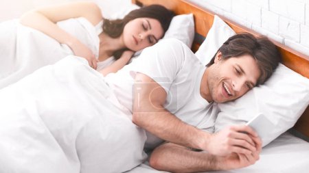 Ein Mann liegt im Bett neben seiner schlafenden Freundin und schaut heimlich auf sein Handy, während sie schläft. Er lächelt verschmitzt und deutet an, dass er sich etwas vorgenommen hat.