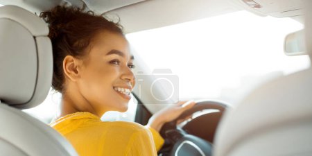 African American young woman with curly brown hair is driving a car. She is smiling and looking out the window. The sun is shining brightly, and the woman is wearing a yellow shirt.