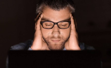 A man wearing glasses concentrates intensely while working on his laptop late at night. His hands are on his head, indicating deep thought or stress.
