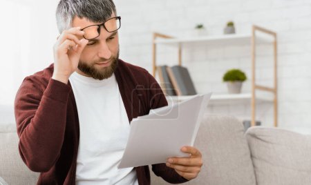 Ein bärtiger Mann mit Brille und kastanienbrauner Strickjacke liest sorgfältig Dokumente durch, während er auf einer hellen Couch in einem hellen, modernen Wohnzimmer sitzt..