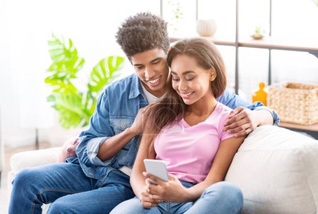 Ein Teenie-Paar sitzt zusammen auf einer Couch und blickt auf ein Smartphone in der Hand der Mädchen. Sie lächeln beide und scheinen glücklich, zusammen zu sein.