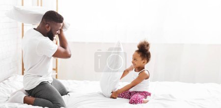 El padre afroamericano y su hija están teniendo una juguetona pelea de almohadas en su dormitorio. Ambos llevan ropa casual y se ven felices y relajados..
