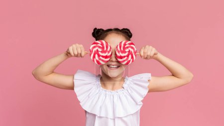 Little sweet tooth. Adorable girl covering her eyes with two colorful lollipops, pink panorama background