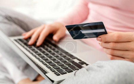 Recortado de mujer se sienta en un escritorio, sosteniendo una tarjeta de crédito en una mano mientras que mecanografía en un ordenador portátil con la otra mano, centrado en introducir la información en el ordenador, compras en línea