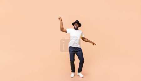 Stylish young African American guy wearing hat dancing, having fun over beige studio background, panorama with copy space