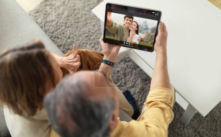 An older couple, a man and a woman, are sitting on a couch and video chatting with a younger couple on a tablet. The older couple is smiling and appears happy to be connected with their loved ones.
