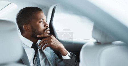 African American businessman in a grey suit, white shirt, and patterned tie sits in the back seat of a car. He is looking out the window and appears to be deep in thought, copy space