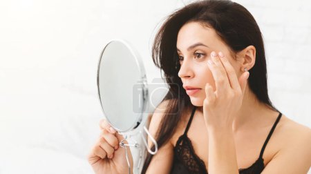 Una mujer está de pie frente a un espejo, observando de cerca su reflejo. Ella aparece enfocada e intencionada mientras examina sus rasgos faciales y expresiones de cerca..