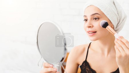 Una mujer está usando un espejo para aplicar maquillaje con un cepillo en una habitación soleada. Lleva una bata de baño y una toalla en la cabeza, lo que sugiere que forma parte de su rutina matutina..