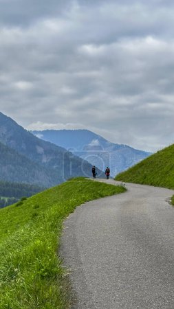 Photo for Santa Maddalena, Val di Funes. Serpentine hill road winds through vibrant greens, two hikers at its end-a glimpse of Alpine beauty in the heart of nature - Royalty Free Image
