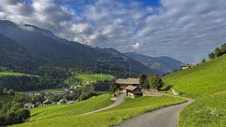 Photo for Santa Maddalena, Val di Funes,A house nestled along a winding road, surrounded by lush pastures, under a clear blue sky with fluffy clouds, framed by the majestic Dolomites-an Alpine haven - Royalty Free Image