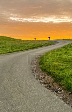 Photo for Hikers, silhouetted against the vibrant hues of the setting sun, manoeuvre a winding road amidst serene landscapes. An action snapshot captured perfectly - Royalty Free Image