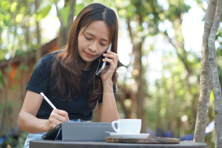 Foto de Mujer asiática mirando las ganancias en su teléfono en una cafetería donde puede ir a trabajar. en medio de la naturaleza verde - Imagen libre de derechos