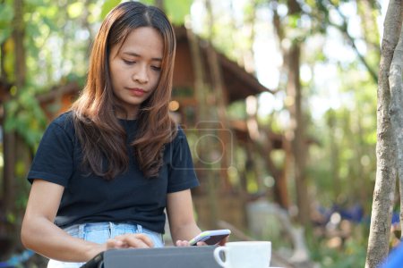 Foto de Mujer asiática mirando las ganancias en su teléfono en una cafetería donde puede ir a trabajar. en medio de la naturaleza verde - Imagen libre de derechos