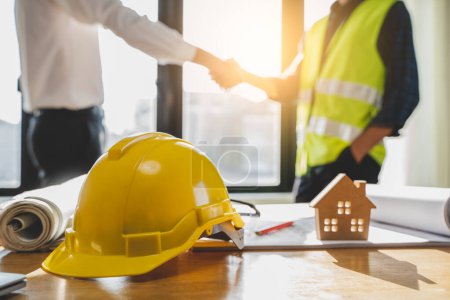 Foto de Close up yellow safety helmet on the table and contractor and customer shaking hands after confirm to build home. - Imagen libre de derechos