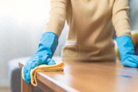 Foto de Trabajadora de servicio ama de llaves feliz limpiando la superficie de la mesa por producto más limpio para limpiar el polvo. - Imagen libre de derechos
