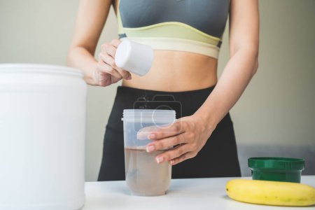 Téléchargez les photos : Young sporty woman pouring protein powder into a cup to make replacement food meal after workout - en image libre de droit