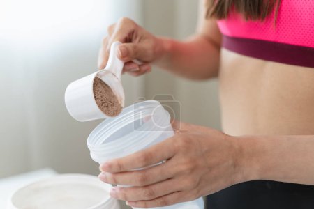 Téléchargez les photos : Close up spoon during young sporty woman pouring protein powder into a cup to make replacement food meal after workout - en image libre de droit