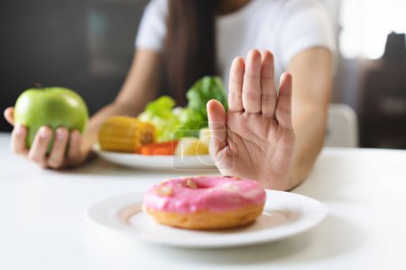 Photo for Woman on dieting for good health concept. Close up female using hand push out her favourite donut and choose green apple and vegetables for good health. - Royalty Free Image