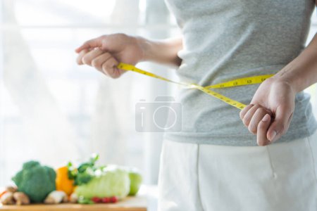 Essen Sie gutes Essen für gute Kondition. Frauen, die ihren Körper mit Maßband messen, haben als Hintergrund ein Gemüse auf dem Tisch. Mädchen überprüft ihre Taillengröße nach unten, um Diät-Ergebnis zu verfolgen.