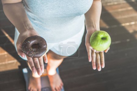 Photo for Choose a right choice for good health. Women is dieting comparison choice between donut and red apple during measuring weight on digital weight measure machine. - Royalty Free Image