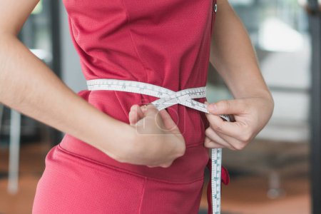 Young slim woman measuring her waist by measure tape after a diet with accessory in sporty gym as background.