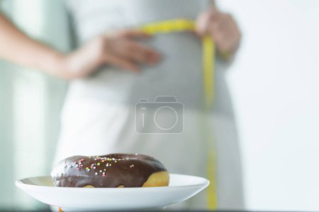 Foto de Dietético para el concepto de forma delgada. Donut en la mesa tiene una mujer midiendo su cintura como fondo. - Imagen libre de derechos