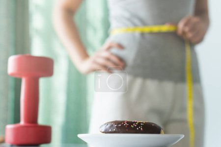 Photo for Dietary for slim shape concept. Donut on the table have a woman measuring her waist as background. - Royalty Free Image