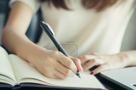 Close-up hand of young female student taking note in to diary book. Selective focus on hand holding a pen.