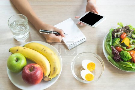 Foto de Dieting and calories control for wellness. Woman using smartphone calculate calories of food in breakfast during dieting for lose weight program and take notes. - Imagen libre de derechos