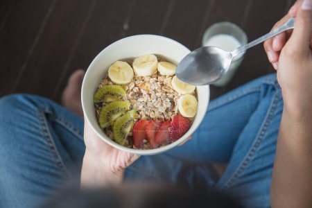 Téléchargez les photos : Eating healthy breakfast concept. Woman holding bowl of cereal and granola and variety fruits and glass of milk on th floor in the morning at home. - en image libre de droit