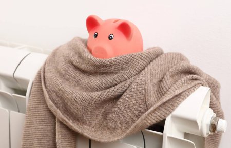 Photo for Cold home, Global energy crisis concept. Price for electricity and heating rising. Piggy bank covered with scarf on radiator - Royalty Free Image
