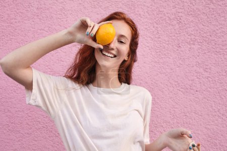 Foto de Retrato de la mujer alegre y despreocupada que esconde su ojo detrás de la naranja mientras posa en la calle sobre el fondo rosado de la pared - Imagen libre de derechos