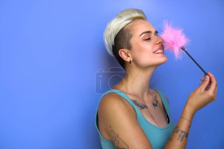 Overjoyed tattooed woman laughing with pink feather stick from sex shop on violet colour background. Erotic toys concept. Stock photo